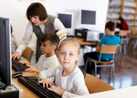 Focused preteen girl studying in computer class of school library on background with classmates and teacherの写真素材