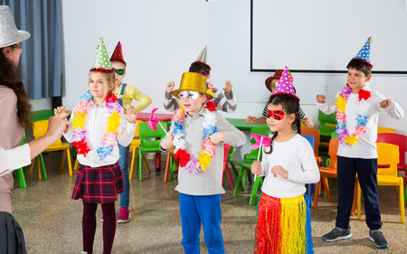 Happy kids and female teacher in funny hats and festive accessories dancing together in schoolroomの写真素材