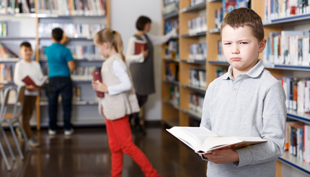 Portrait of upset tween boy browsing book in school libraryの写真素材