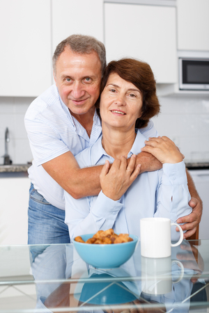 Portrait of happy mature couple hugging near kitchen table before drinking teaの写真素材