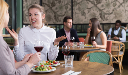 Cheerful young woman with female friend enjoying dinner and drinking red wine in restaurantの写真素材