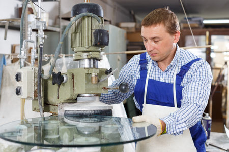 Confident male glazier with female assistant working on curved glass beveling machine in workroom