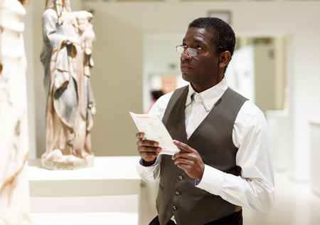 Afro man wearing glasses visiting exposition of museum and reading guidebookの写真素材