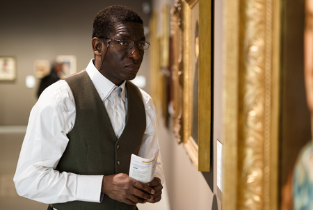 Positive Afro-American man with booklet standing at hall of museum and looking at paintingの写真素材