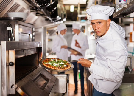 Portrait of confident chef working in modern restaurant kitchen, getting ready pizza out of ovenの写真素材