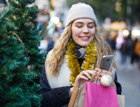 Portrait of happy girl in knitted hat and scarf talking on smartphone at Christmas trees fairの写真素材