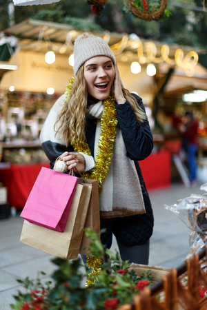 Cheerful girl in knitted hat and scarf with paper bags posing near Christmas fair shopsの写真素材