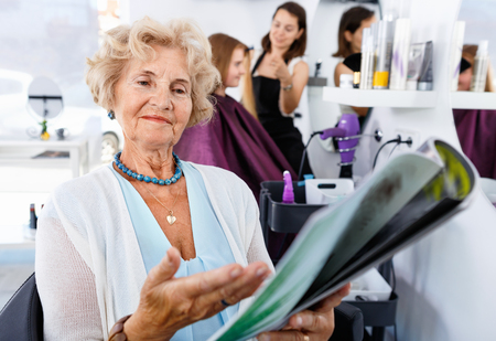 Smiling cheerful  elderly female client sitting with magazine in chair in hair salon, choosing new hairstyleの写真素材