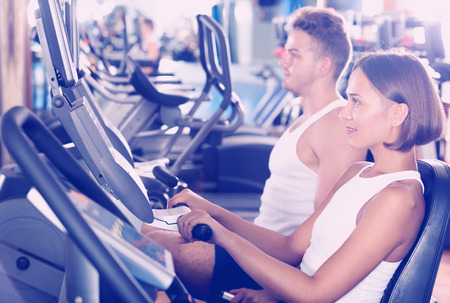 Glad man and young woman warming up before workout using cycling cardio machinesの写真素材