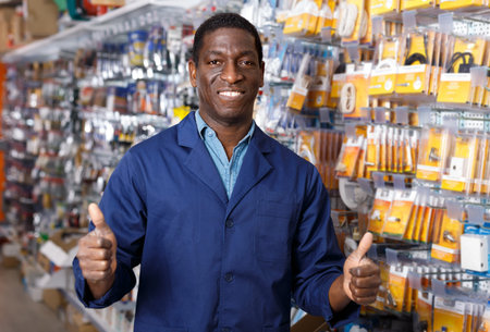 Adult African American seller of household store standing near racks with goods, giving thumbs up