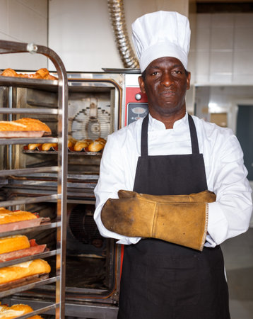 Confident worker of bakery wearing uniform and heat resistant gloves standing with arms folded near...