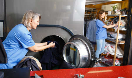 Efficient glad woman in uniform taking out clothes from washing machine at laundryの写真素材