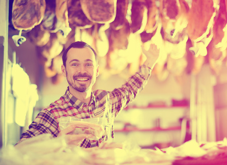 Smiling man seller showing sorts of meat in butcherâs shopの写真素材
