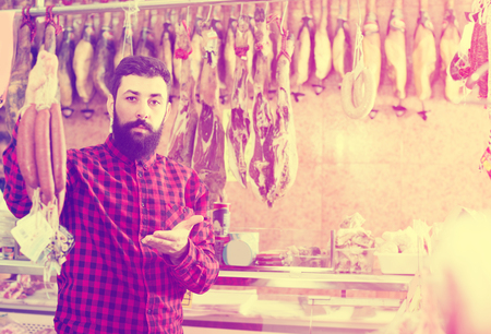 Smiling young guy deciding on best sausage in butcherâs shopの写真素材