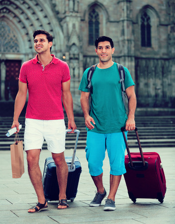 Happy positive smiling men tourists are walking with suitcases in unknown city.の写真素材