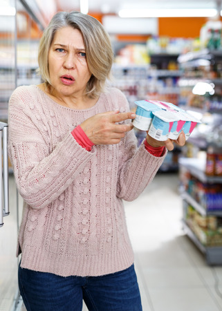 Senior woman choosing necessary foodstuffs in grocery store, looking unpleasantly surprised by reading product contentsの写真素材