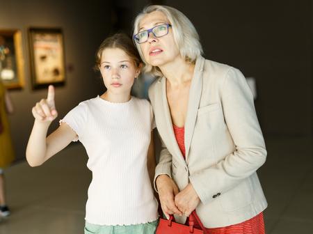 Attentive tween girl with senior woman looking with interest at gallery expositionの写真素材