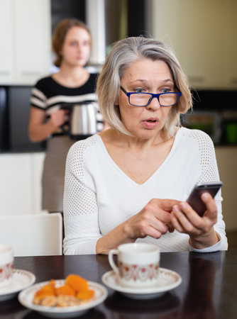 Upset mature mother using smartphone in kitchen interior while adult daughter making teaの写真素材