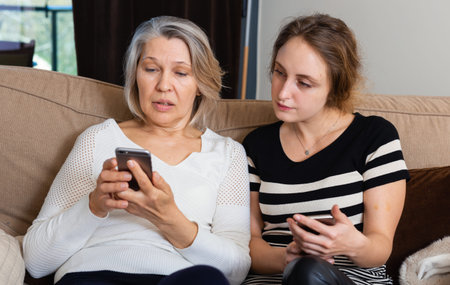 Mature woman and adult daughter using phones sitting on sofa at home