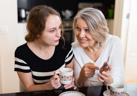 Happy mother and daughter using mobile gadget at kitchen table at homeの写真素材
