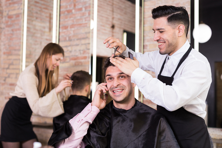 Cheerful man talking on phone while male barber making his hairstyle in barbershopの写真素材