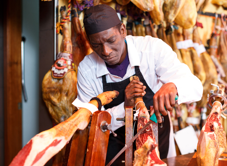 Confident African American salesman slicing jamon fixed on jamonera, preparing for saleの写真素材