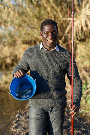 Positive African man standing near river and holding fishing rod and bucket with catchの写真素材