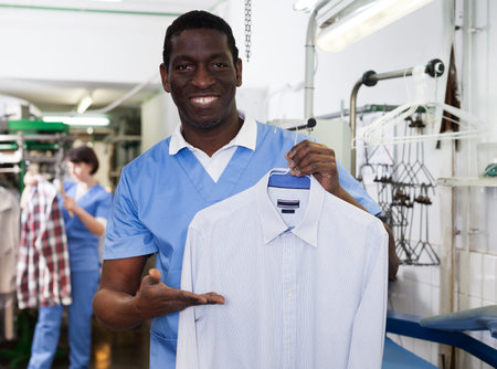 Cheerful African American man worker of dry cleaner showing clean clothes after dry cleaning