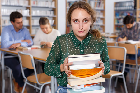 Portrait of adult girl with books in hands sitting in classroom with studying fellow studentsの写真素材