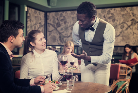 Positive elegant couple talking with polite African American waiter while dining in cozy restaurantの写真素材