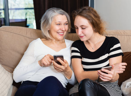 Mature woman and adult daughter using phones sitting on sofa at home
