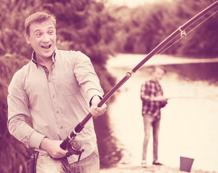 portrait of cheerful smiling man casting line for fishing on riverの写真素材