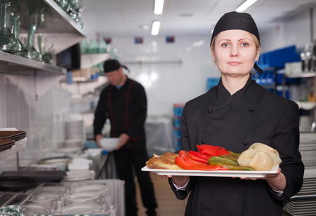 Proud woman cook standing in restaurant kitchen with finished dish, ready to serve it to guestsの写真素材