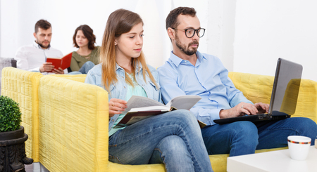 Friendly young cheerful  woman and man relaxing in cozy hostel lobby, spending time with laptop and bookの写真素材