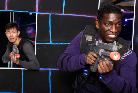 Portrait of emotional African American man aiming laser gun at other players during lasertag game on dark arenaの写真素材