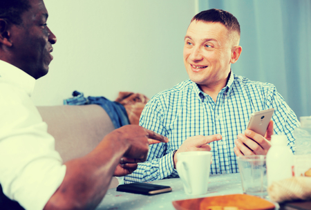 Smiling man meeting with African friend at home, looking at phone and discussingの写真素材