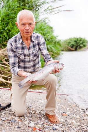 Senior male fisher examining catch fish while angling at lakeside on summer dayの写真素材