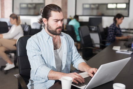 Focused bearded freelancer concentrated on work with laptop in coworking space with international teamの写真素材