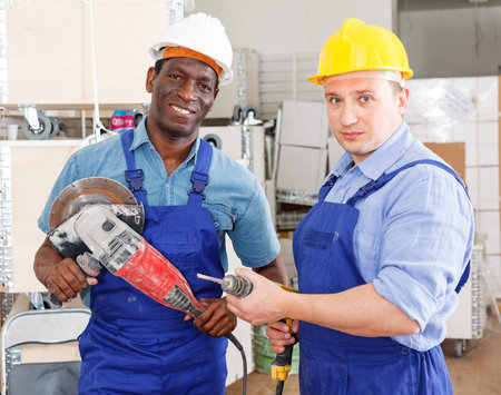 Confident male contractors working with handheld power tools in overhauls indoors
