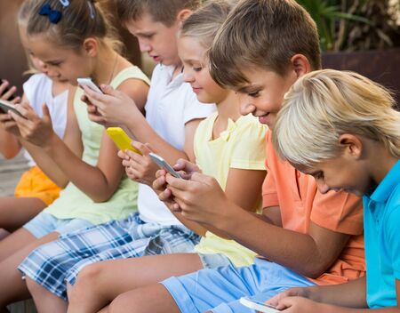 happy american children in school age looking at mobile phones and sitting outdoorsの写真素材