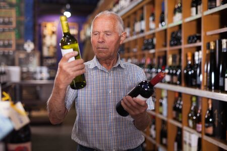 Positive grey-haired elderly man choosing wine in modern wineshopの写真素材