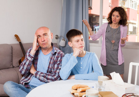 Frowning teenager and adult man listening to reprimanding from outraged woman at home