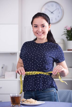 Smiling young woman measuring waist with tape near table with breakfastの写真素材