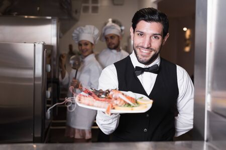 Smiling bearded waiter holding seafood dish in kitchen of fish restaurantの写真素材