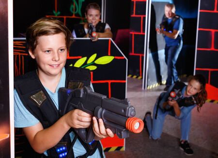 Excited  cheerful positive smiling boy aiming laser gun at other players during laser tag game in dark roomの写真素材