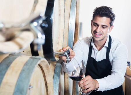 Happy man seller wearing apron pouring wine from wood into glass in cellarの写真素材