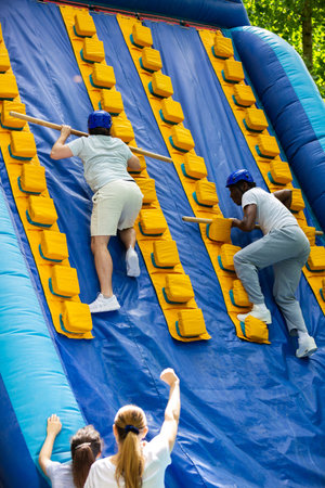Two adult men having funny competition in climbing on inflatable castle with wooden sticks in...