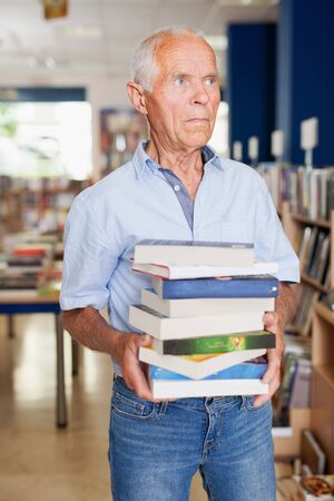 Portrait of senior male with pile of books in hands in library interiorの写真素材