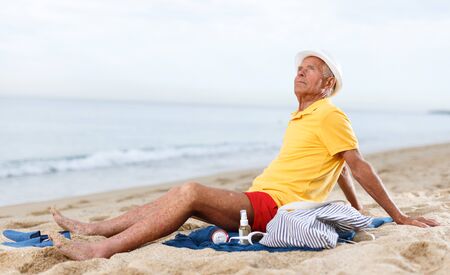 Satisfied glad elderly man  sitting on the sand near the oceanの写真素材