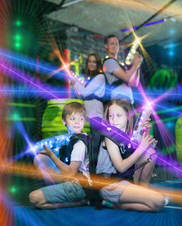 Cheerful little boy and girl sitting back to back on bright beams with laser pistols in dark lasertag room during game with parentsの写真素材